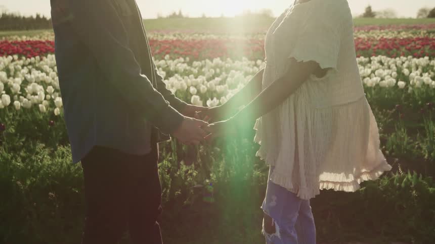 A man and woman face one another and hold hands in front of a field of flowers