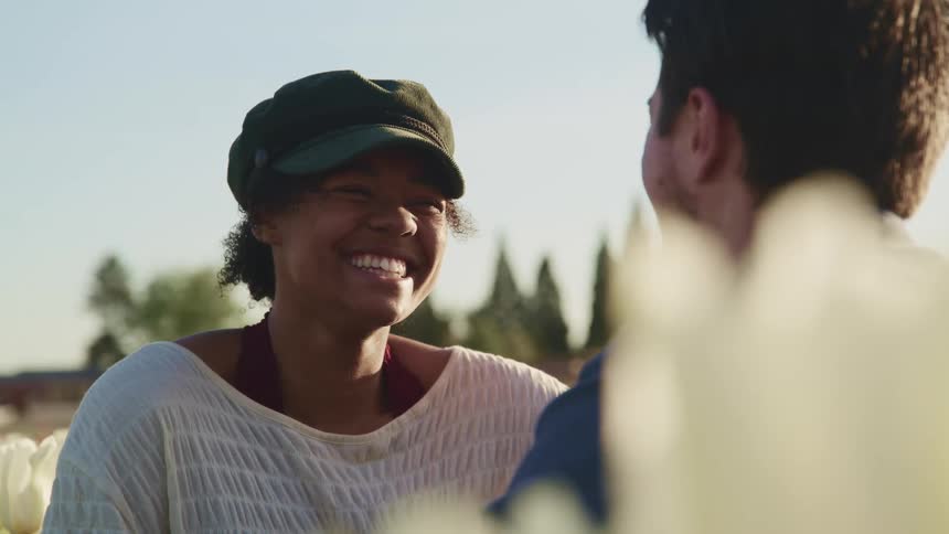 Man and woman smile at each other in field of white flowers with a building and trees on the horizon