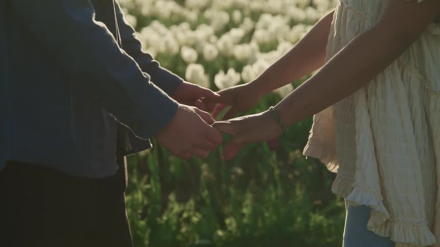A man and woman standing in a field of flowers face one another and hold hands