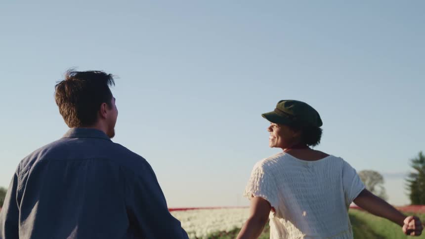 A smiling woman and man hold hands while they gaze at each other and run between rows of flowers in a field
