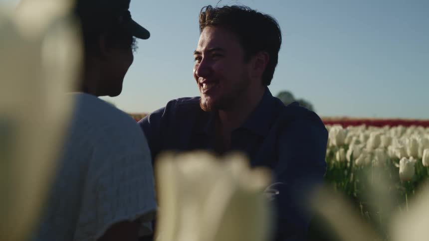Black woman with a white shawl and white man with a beard sit down and look at each other with love in tulip fields