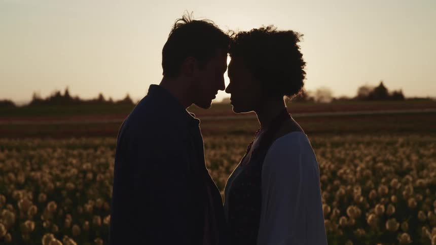 An asian male and a black woman embrace and touch foreheads as they stand in a field of flowers