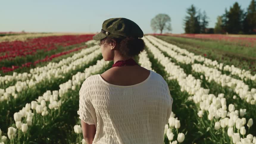 A young woman in a cap stands in vast field of rows of white and red flowers as a gentle breeze rustles her clothing