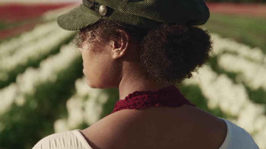 Young woman wearing a hat stands in front of a tulip field at sunset