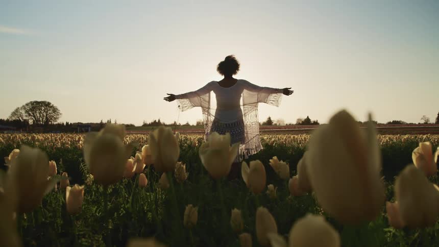 A woman standing in a field of flowers raises her arms to her sides