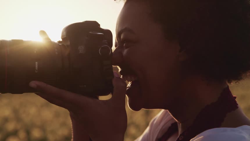 A smiling black female photographer takes photographs in field under a low sun