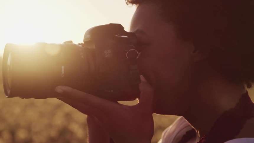 A smiling black female photographer brings a camera to her eye while she stands in a field under a low sun