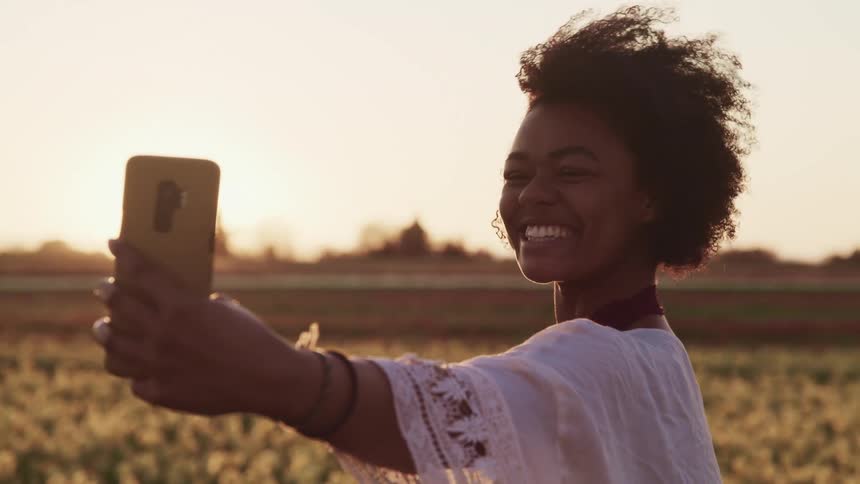 Black woman with curly hair and a white shawl smiles broadly and takes a selfie with a smartphone at sunset in tulip fields