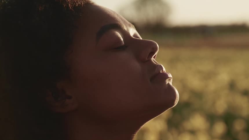 A woman facing upwards with her eyes closed with the sun shining on her face with a field the sky and clouds in the background