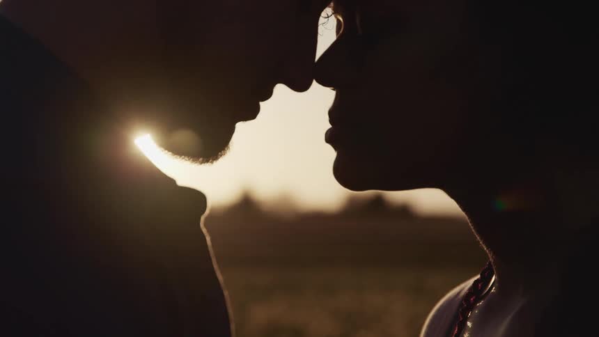 Man and woman standing and touching their noses to each others is silhouetted against the sun in tulip fields