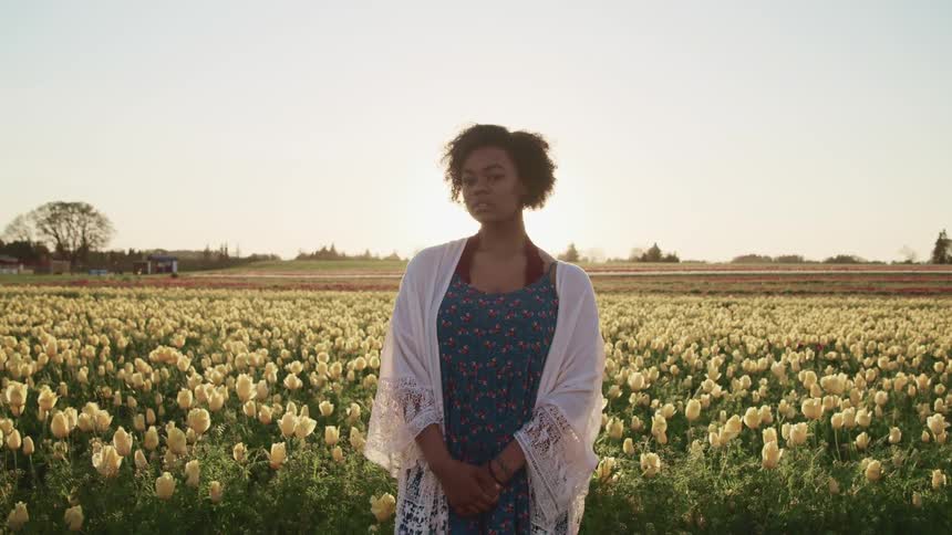 Black woman with curly hair and a white shawl stands in white tulip fields at sunset
