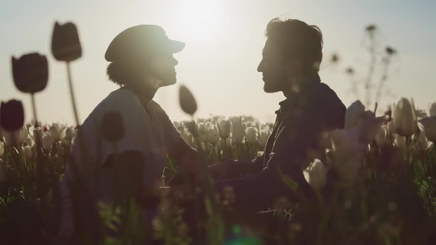 A woman and man sit facing one another in a field of white flowers and look into one anothers eyes while holding hands