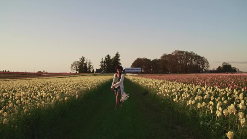 Black woman with curly hair and a white shawl walks happily through tulip fields at sunset
