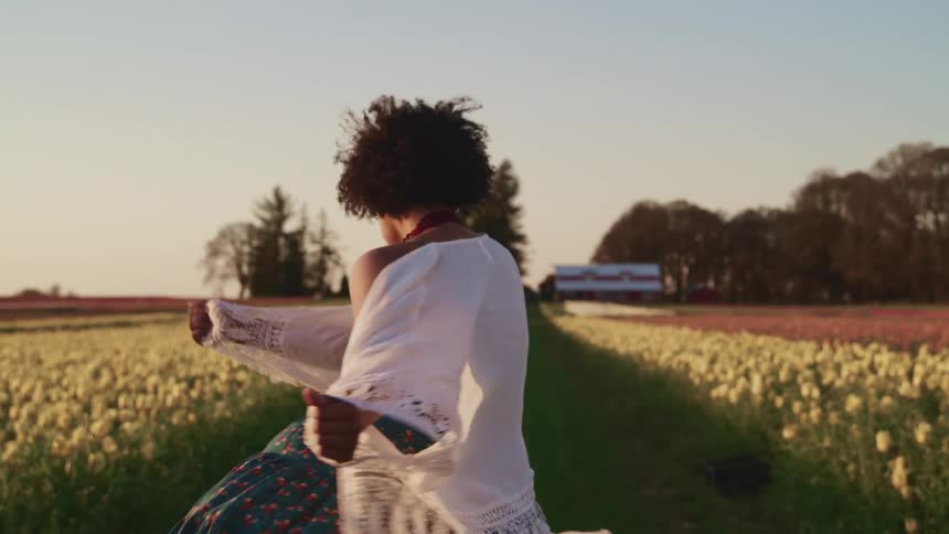 Woman with curly hair and a white shawl walks happily and turns around in tulip fields at sunset
