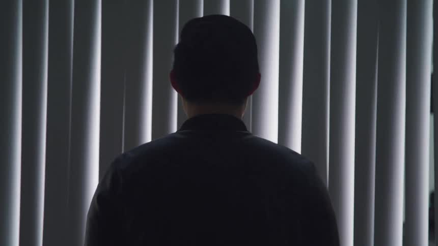 A person stands in front of closed vertical blinds that are shaking side to side very rapidly