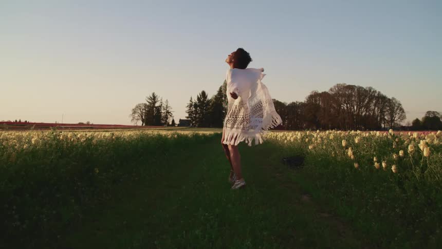 Black woman with curly hair and a white shawl walks happily in tulip fields at sunset