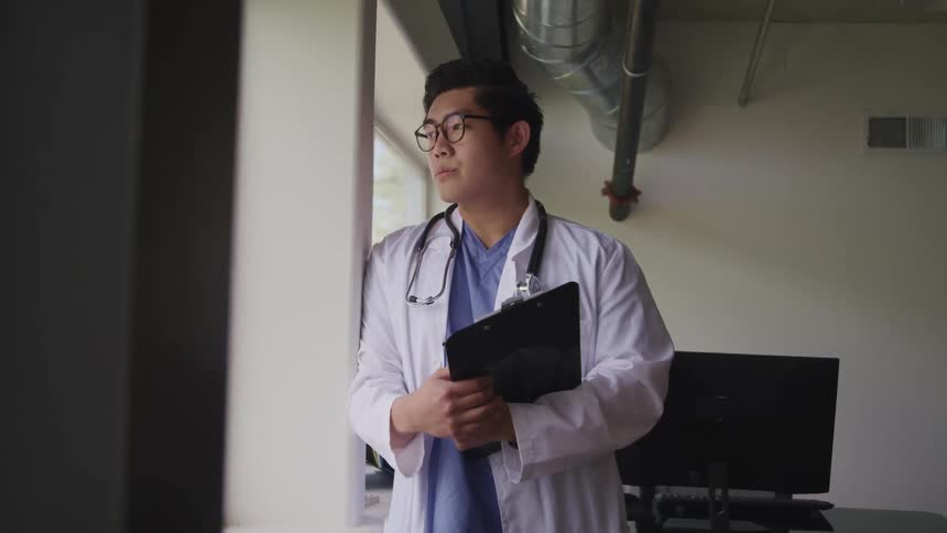A male doctor holds a clipboard and stands in a room looking solemn out the window