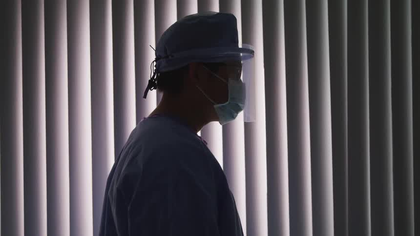 A doctor wears a mask and a face shield as he stands in front of a window