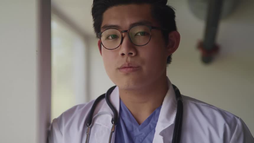 A young asian male physician leans against the wall nearby a window