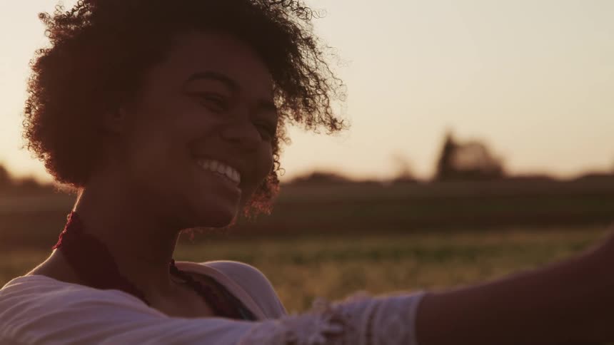 Black woman with curly hair and a white shawl smiles broadly and takes a selfie with a smartphone at sunset in tulip fields