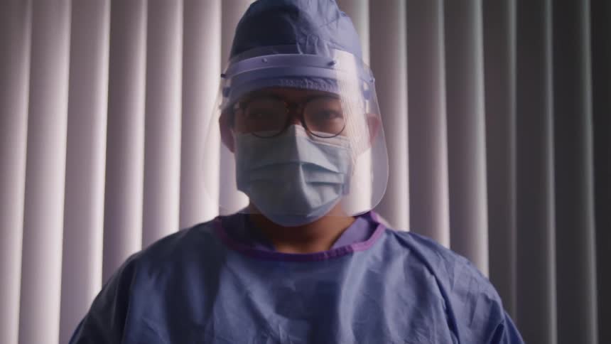Doctor wearing surgical gear including a face shield over his eyeglasses stands in front of blinds