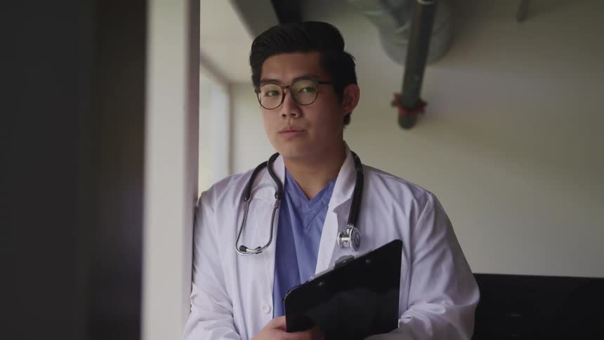 A male doctor stands next to a window holding a clipboard