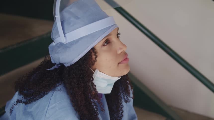 A young woman in surgical scrubs sits on stairs with her face mask under her chin as she relaxes