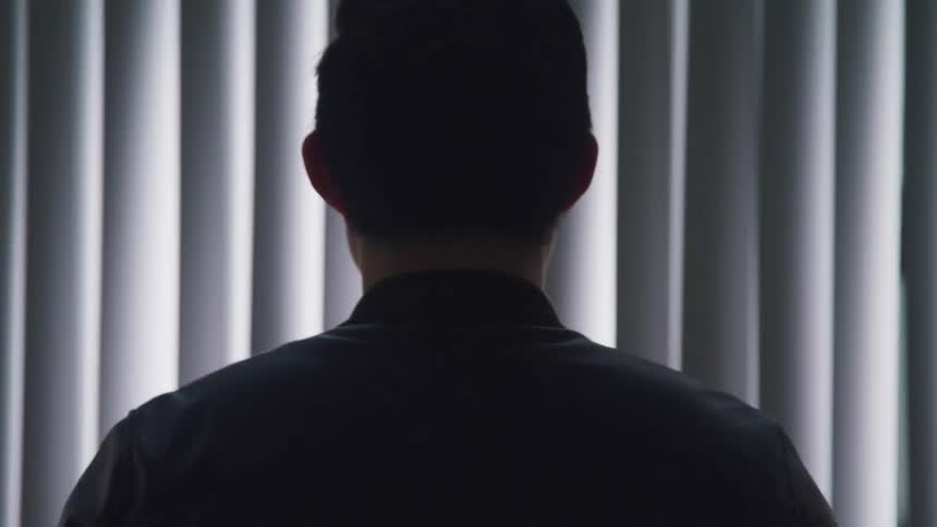 A man standing and facing blinds covering a window