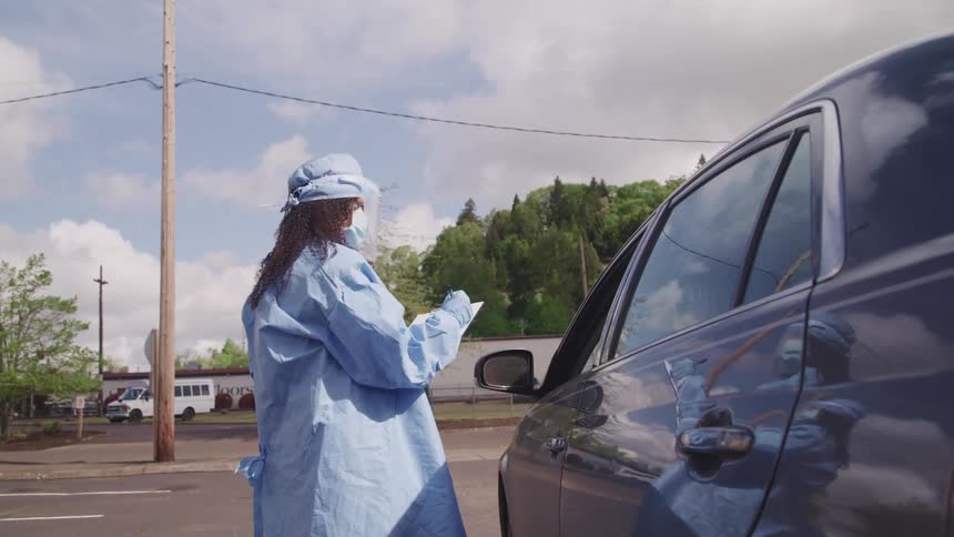 A woman dressed in full medical scrubs and ppe stands beside a vehicle with the drivers side window open