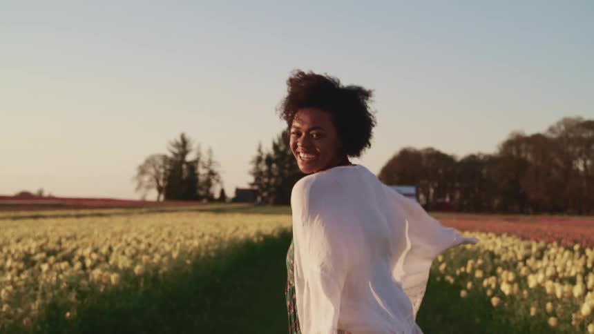 Black woman with curly hair and a white shawl walks happily through tulip fields at sunset
