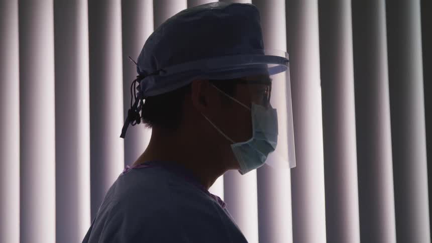 Doctor in scrubs and face mask and face shield stands in front of blinds
