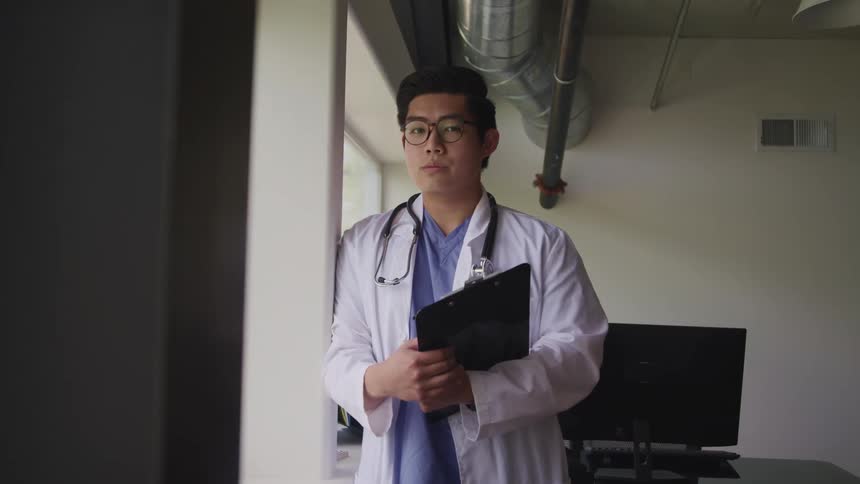 Asian male doctor with glasses and a stethoscope around his neck faces the camera with eye contact in the room