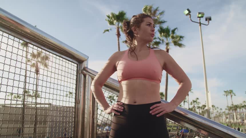 Woman in sport clothes stands next to a metallic fence in a park at sunset