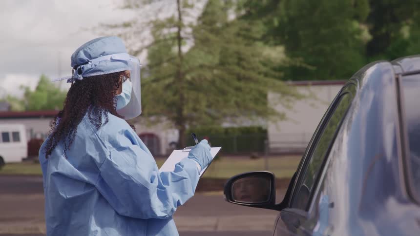 Medical personnel in protective gear talks to male driver in car taking notes on clipboard