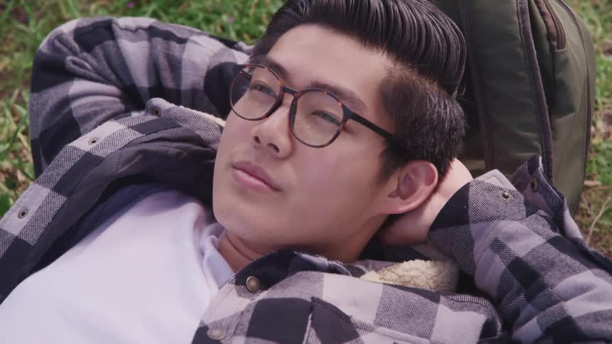 A young asian man rests in the grass with his head on his backpack