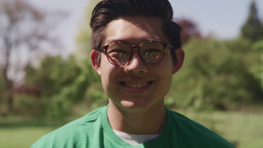 Asian man with a green t shirt and glasses smiles broadly and faces the camera with eye contact in a public park