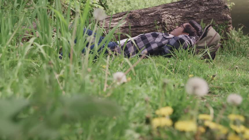 Young man sleeping on the grass near a tree trunk using a backpack as a pillow