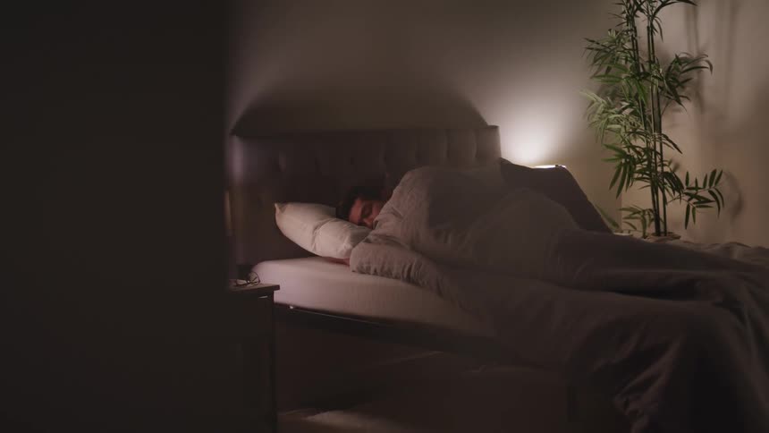 A man sleeps on a gray upholstered bed with a gray comforter pulled up to his chin
