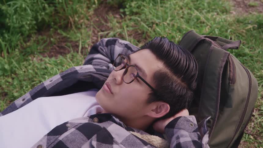 Young asian man wearing glasses lays in the grass using backpack as pillow