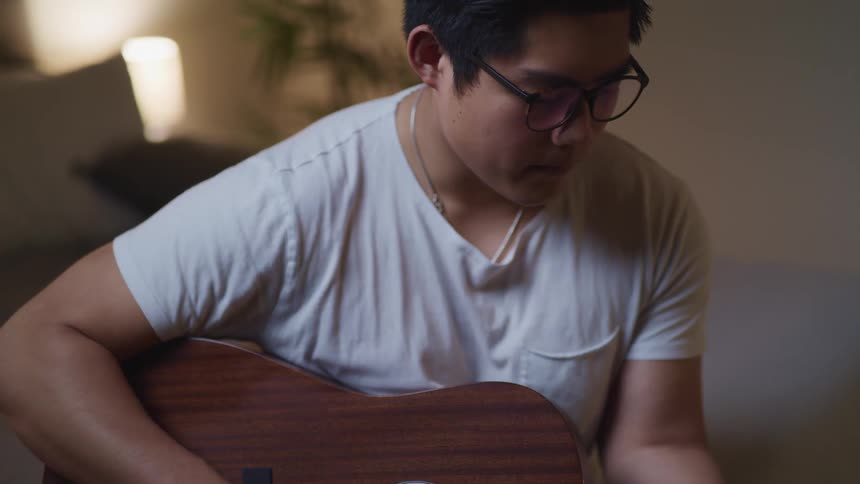 Asian man with a white t shirt and glasses plays a guitar