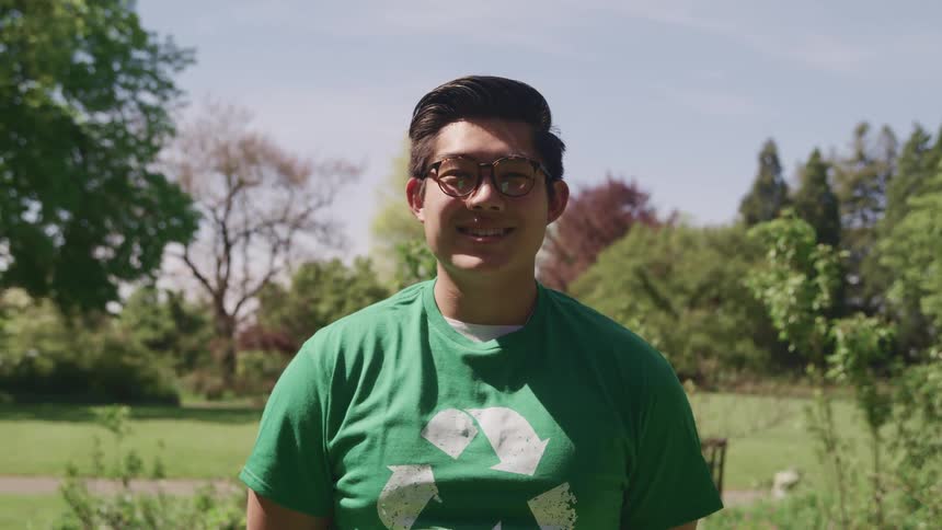 Asian chubby man with a green t shirt stands and faces the camera with eye contact in a public park