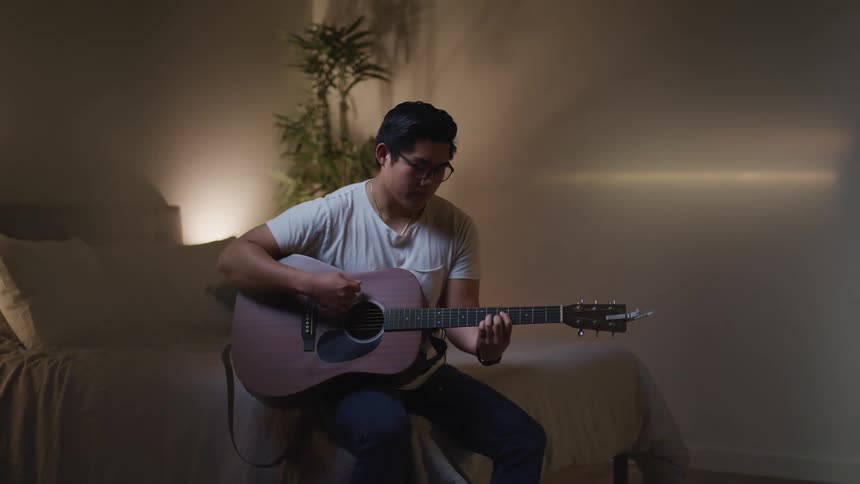 A teenage boy sits on a bed and plays the guitar