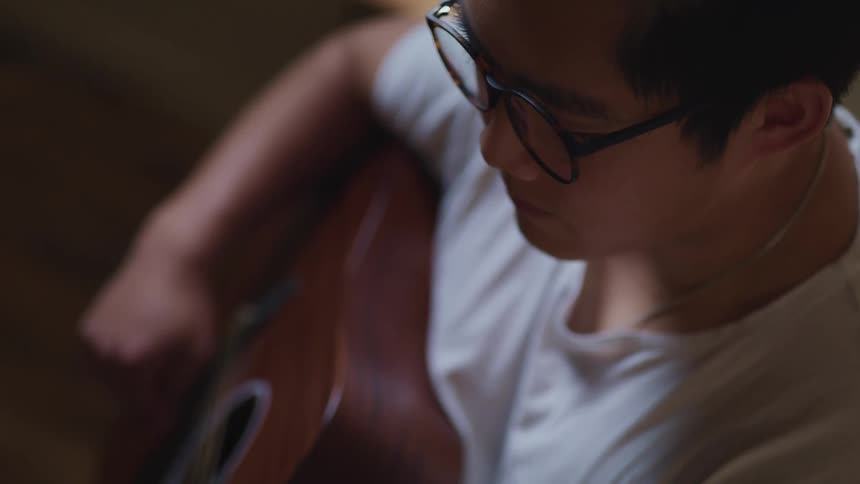 Asian man with a white t shirt and glasses plays a guitar