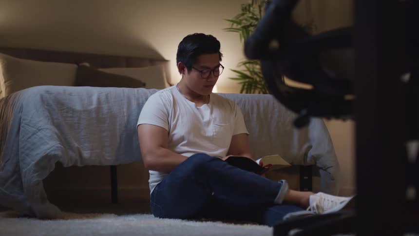 Male sitting on floor at the end of the bed in a bedroom turns the page in the book he is reading