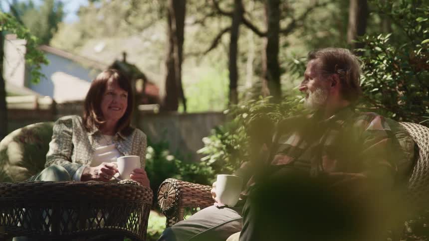 Man with a grey beard a pink striped shirt and woman with brown straight hair sit in armchairs and talk to each other in the garden