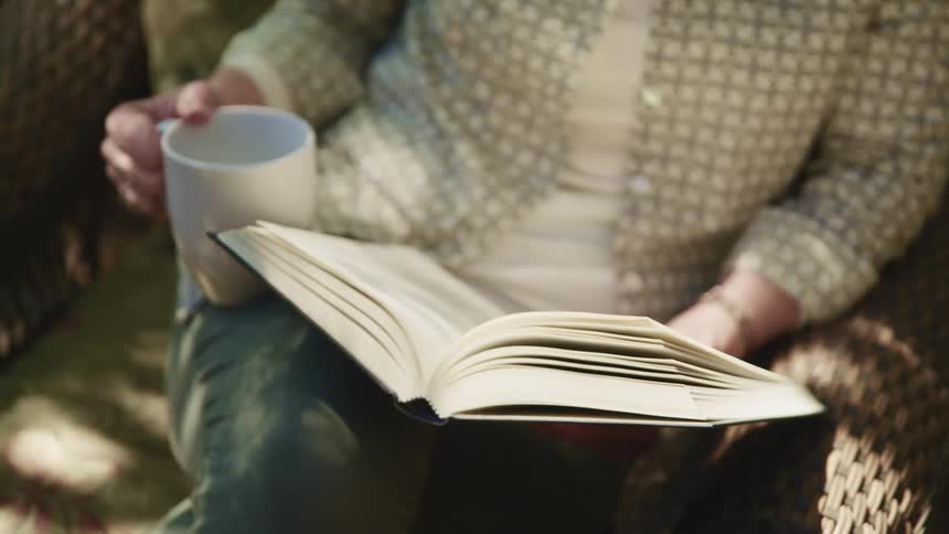 A woman reads a book while sitting outside in a wicker chair
