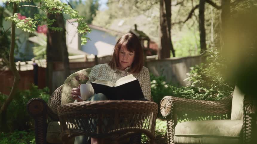 Woman sitting in the garden reads a book while holding a cup
