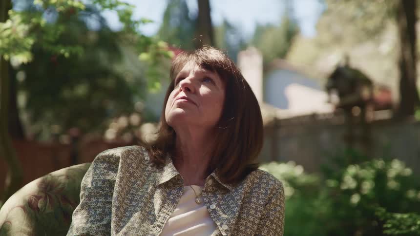 A woman sitting on a chair in a backyard with plants trees and a fence in the background