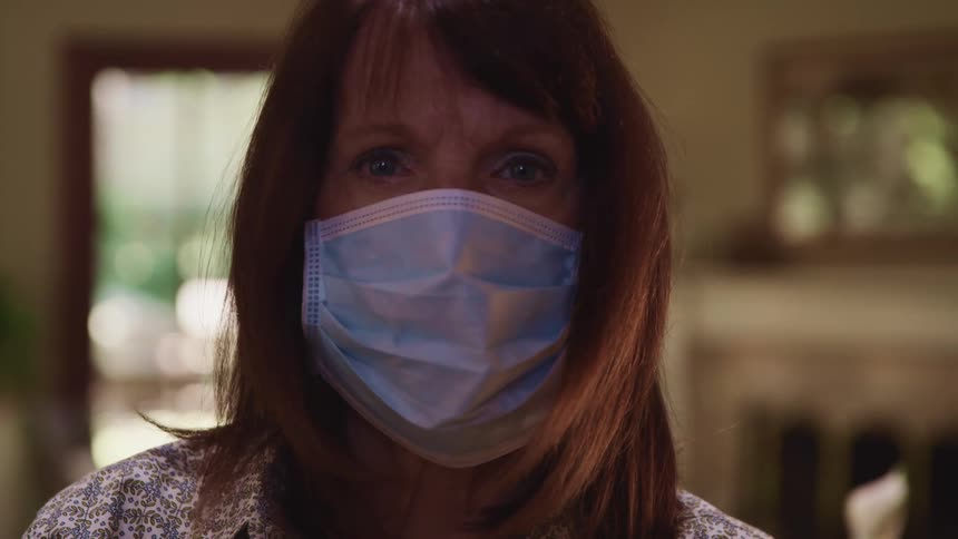 Middle aged woman with brown hair and a light blue mask raises her head and faces the camera with eye contact