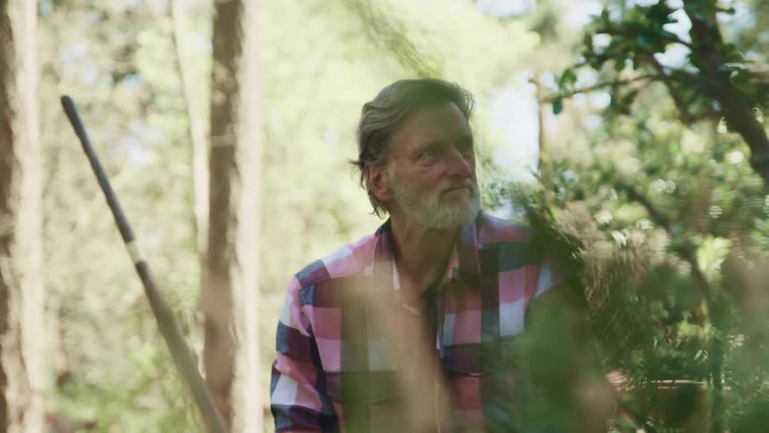 An elderly man sits outside surrounded by trees and looks off in to the distance